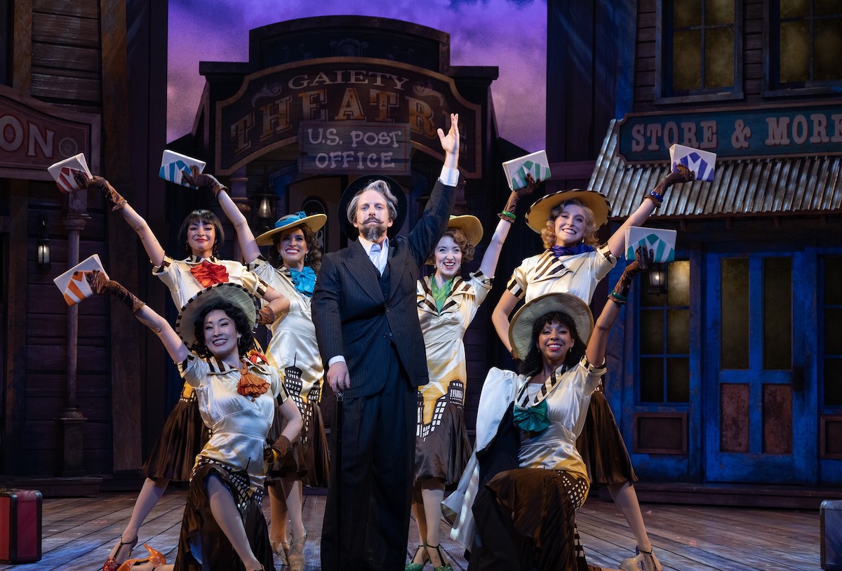 An actor with a fancy mustache poses between six women in front of a historic town set.