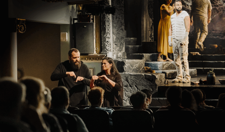 photograph focused on two sign language translators in the mid-ground with out of focus audience members in the foreground and three actors in costume at stage right in the background