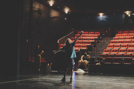 Shot from behind, a young actor stands with outstretched arms in front of empty auditorium seats as two people watch.