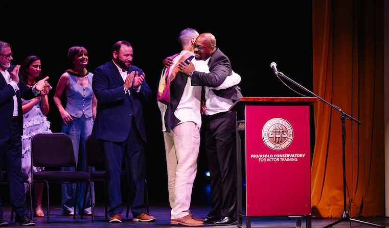 Two men embracing next to a podium on a stage with onlookers applauding behind them.